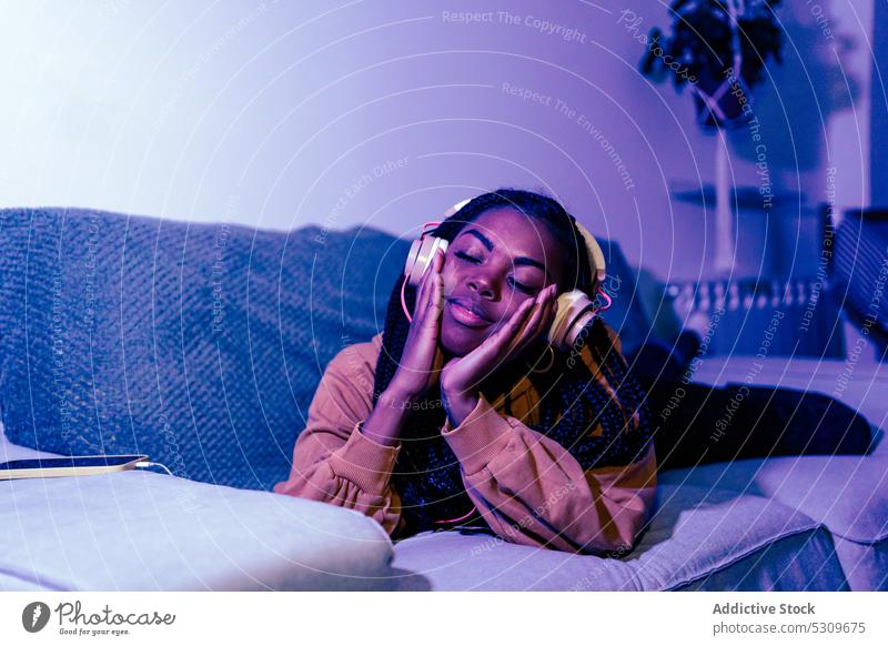 Relaxed black woman listening to music with closed eyes on sofa headphones neon lying home relax calm song eyes closed light young ethnic device sound gadget