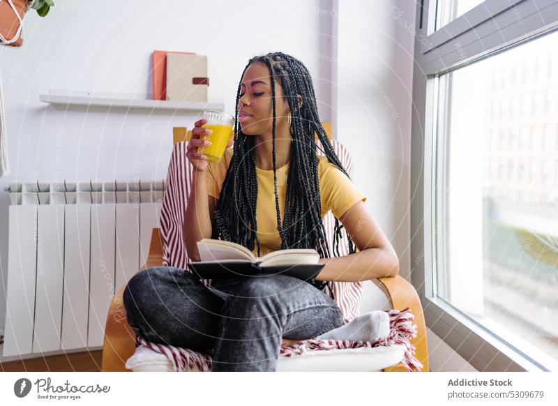 Calm black woman with book drinking orange juice read armchair rest content home literature female young african american ethnic beverage comfort window enjoy