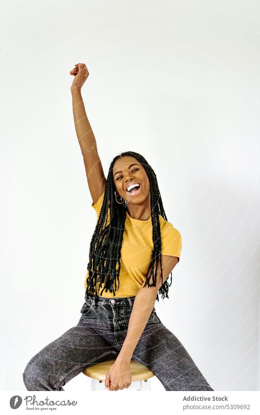 Smiling black woman sitting on chair against white wall cheerful smile positive trendy arm raised braid content happy appearance style confident female delight