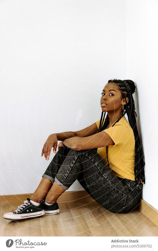 Confident black woman sitting on floor and looking at camera calm trendy serious style appearance wall confident outfit fashion female young african american