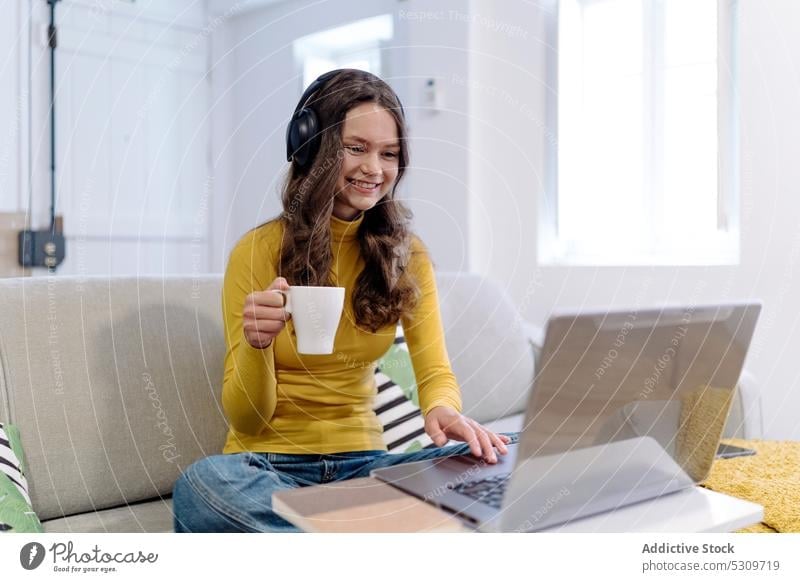 Focused woman in headphones with coffee cup working on laptop on sofa remote freelance using telework browsing young home female online device internet gadget
