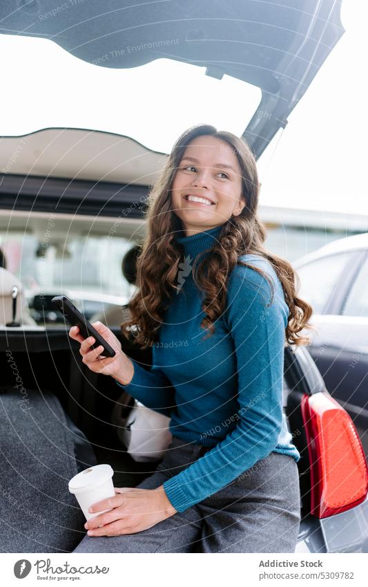 Smiling woman on smartphone sitting in car trunk smile automobile happy coffee female browsing takeaway young gadget long hair device connection online