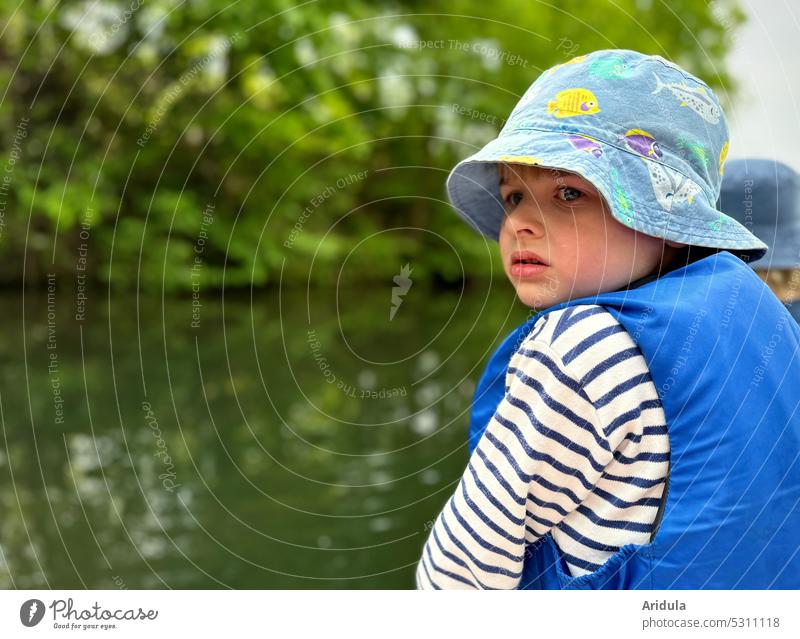 Child sits in a boat and looks skeptically backwards at the water. It wears a sun hat and a life jacket Water Pedalo Looking Skeptical Insecure Fear anxiously