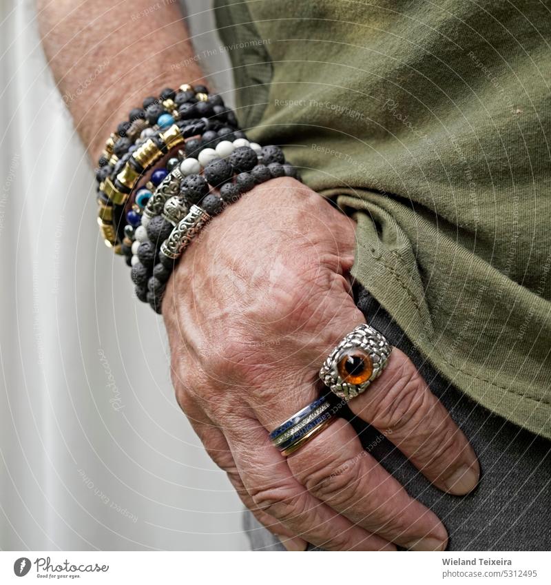 An older man puts his hand in the pocket of his gray jeans. He wears lava stone and other breaded bracelets and special rings. person Stone Black Design