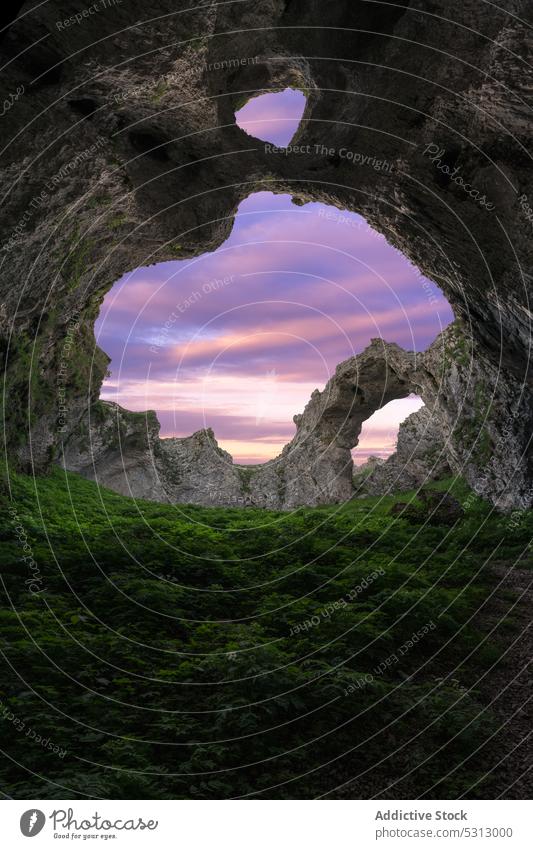 Double hole cave in mountainous terrain against sunset sky rocky formation green grass colorful picturesque sundown landscape scenery vivid vibrant breathtaking