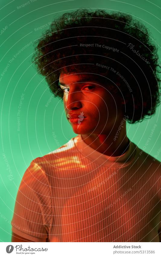 Hispanic guy standing against colorful illumination looking at camera man model multicolored afro hair illuminate projector glow creative studio shot young male