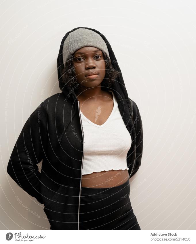 Calm black woman in hat looking at camera serious confident wall outfit style cool hoodie portrait young appearance calm personality female individuality casual