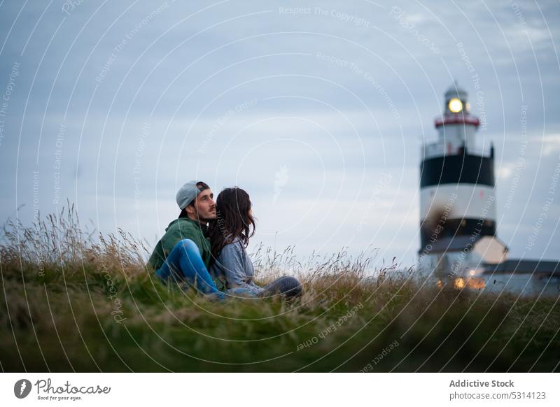 Young couple sitting on hill with lighthouse traveler countryside nature highland relax grass journey ireland weekend tourist meadow romantic vacation ocean