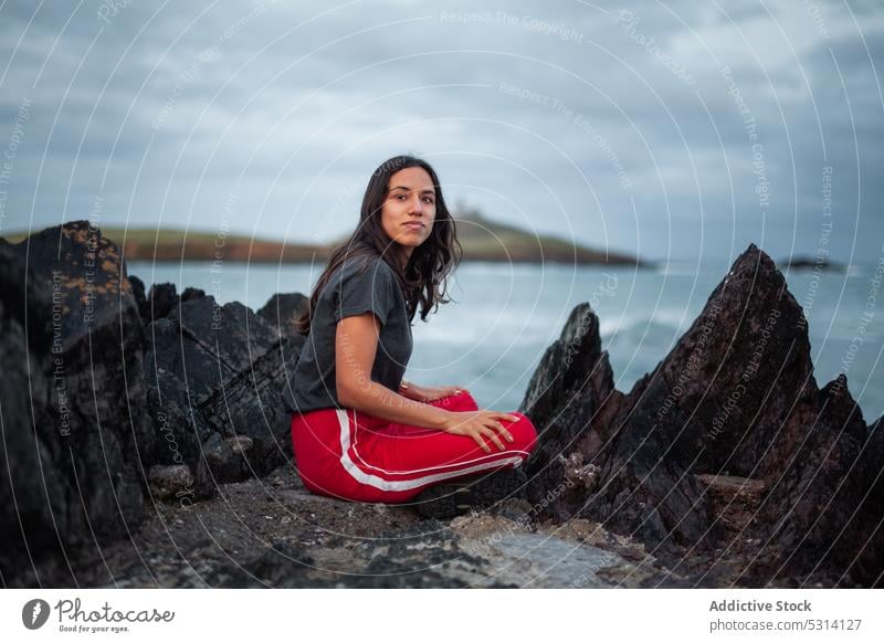 Thoughtful young woman sitting on rock near ocean cliff sea coast mountain nature rocky foam shore ireland sky happy wave female cloudy water positive admire