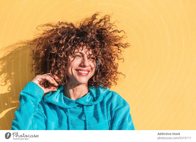 Charming curly haired woman touching hair against yellow wall portrait style individuality color cheerful touch hair charming outfit female glad smile