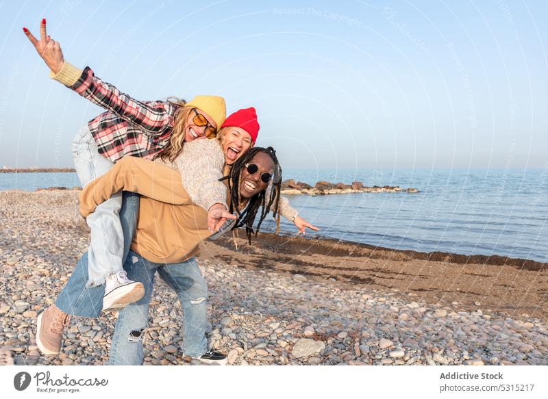 Happy group of diverse friends having fun on beach near sea man women piggyback v sign coast gesture two fingers smile together carry couple peace summer