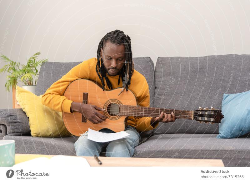 Serious African American man with dreadlocks looking at paper with music notes musician guitar hobby instrument guitarist living room player sit melody talent