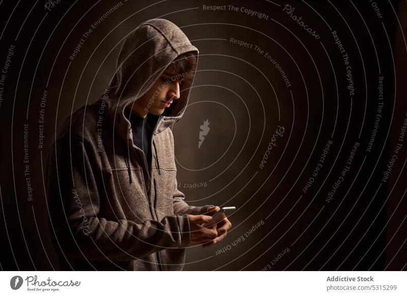 Concentrated man in hood browsing smartphone in dark studio using message hoodie focus serious gadget mobile device male concentrate internet communicate young