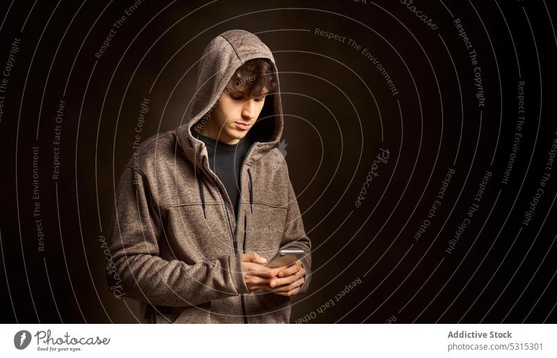 Concentrated man in hood browsing smartphone in dark studio using message hoodie focus serious gadget mobile device male concentrate internet communicate young
