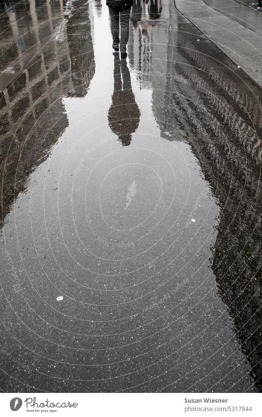 A person (half) with reflection (whole) walks on a wet street through the urban canyons of a big city. after the rain Reflections in the water City