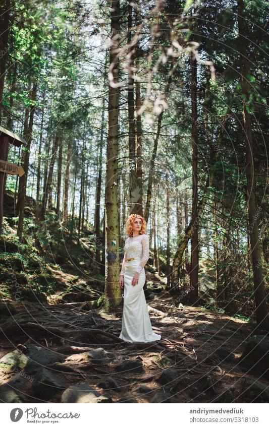 beautiful young red-haired bride in the forest with a floral wreath on her head. woman in long white dress outdoors on summer day. wedding day woods bouquet