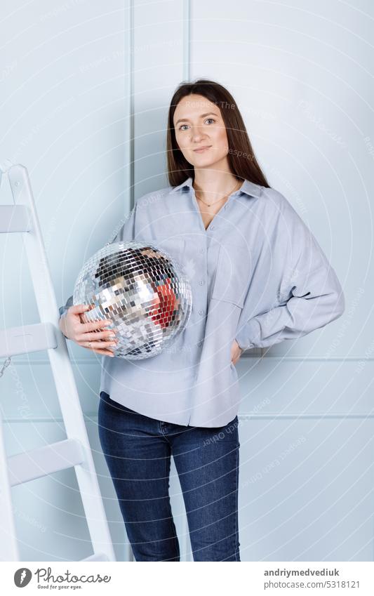 beautiful young woman with a disco ball in her hands. girl with long hair in blue clothes near stepladder on blue wall background cute lovely fashion party