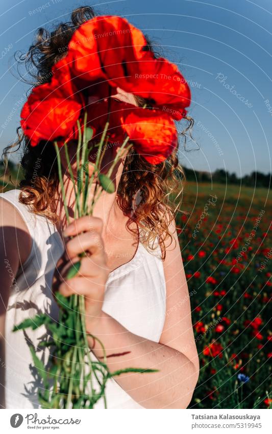 Selfie of a curly red-haired woman covering her face with a bunch of red field poppies among a poppy field. selfie wildflowers bouquet sunny day sunlight female