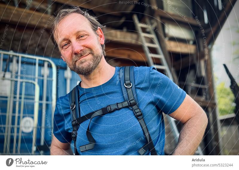 Man in blue t-shirt and with backpack looks friendly into camera, storage shed/workshop in background kind portrait Face Smiling Human being Looking Facial hair