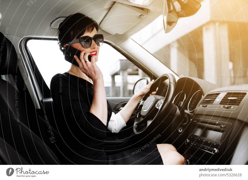 Attractive successful business woman sits inside of the luxury car and talking on her smartphone. Business owner woman in sunglasses driving a car solves work issues on the way to office. Lifestyle