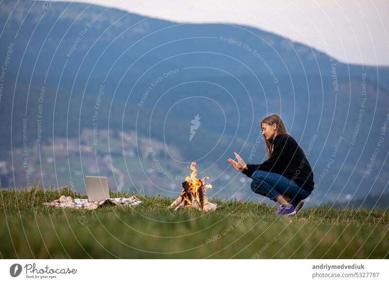 young woman freelancer sits by the fire and warms his hands in the mountains in the evening. tourist girl resting and working on laptop outdoor notebook travel