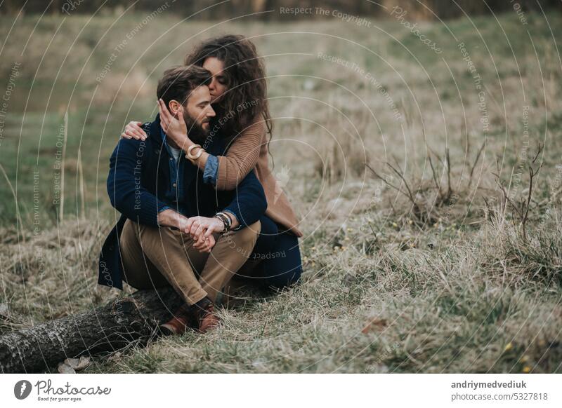 Happy caucasian lovers are sitting on the shore of the lake. Young couple is hugging on autumn day outdoors. A bearded man and curly woman in love. Valentine's Day. Concept of love and family.