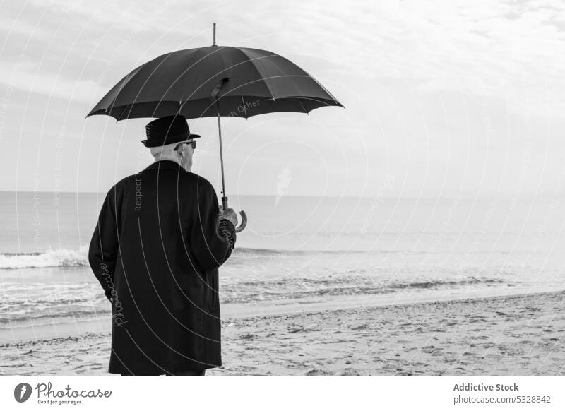 Unrecognizable matured man in cozy suit with umbrella on sandy beach sea shore lonely solitude ocean wave melancholy coast warm clothes senior seaside nature