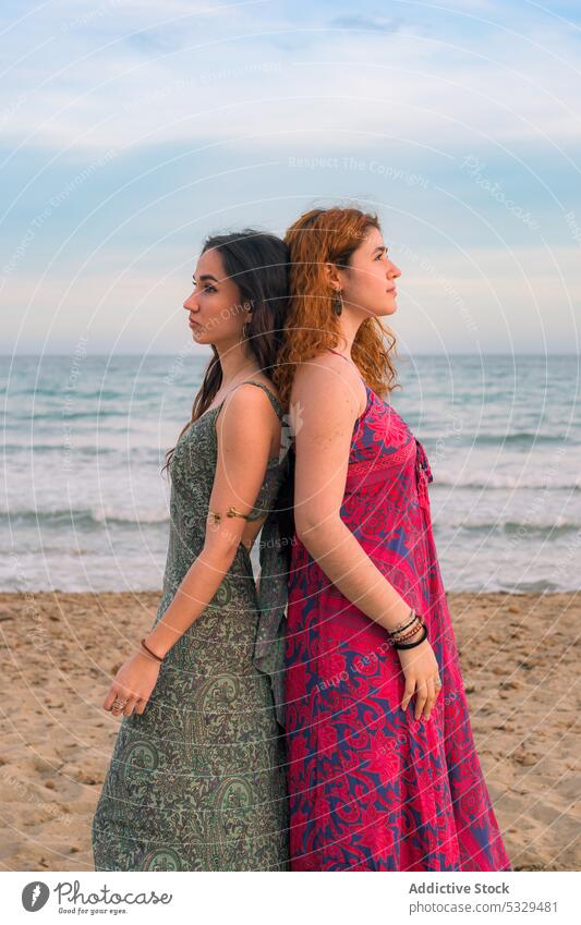 Young women in dresses standing on beach dreamy thoughtful sea wave girlfriend back to back summer ocean together shore coast style young female water holiday