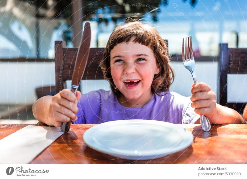 Funny little girl with silverware and plate in cafe kid mouth opened child wait table hungry childhood food wooden cute preschool adorable sweet order serve