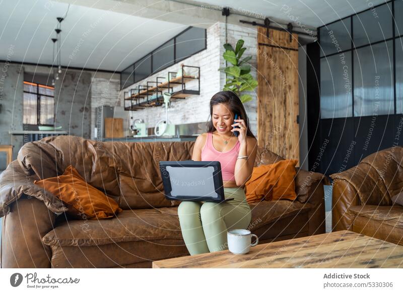 Asian woman talking on smartphone while working on laptop freelance remote home sofa call gadget conversation female young casual project connection phone call