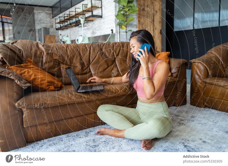 Cheerful woman browsing laptop on sofa while making calls on floor smartphone freelance cozy barefoot apartment living room rest home gadget weekend using