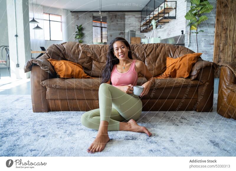 Relaxed woman drinking coffee sitting on the floor in cozy living room rest asian smile sofa hot drink home comfort relax happy young cup lifestyle female couch
