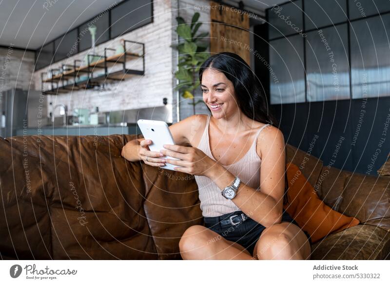 Smiling young woman using tablet on sofa smartphone home smile cheerful cozy couch living room gadget device rest casual female online mobile internet surfing