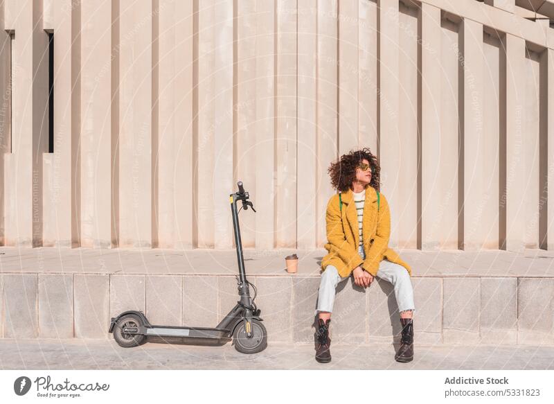 Calm woman sitting near electric scooter on street coffee takeaway trendy urban style ledge modern hipster female young transport curly hair to go city