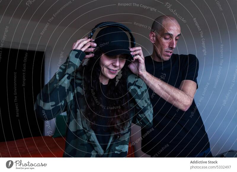 Man putting headphones on woman at home couple console listen music dj put on together casual sweatshirt smile adult style bald daylight long hair cap