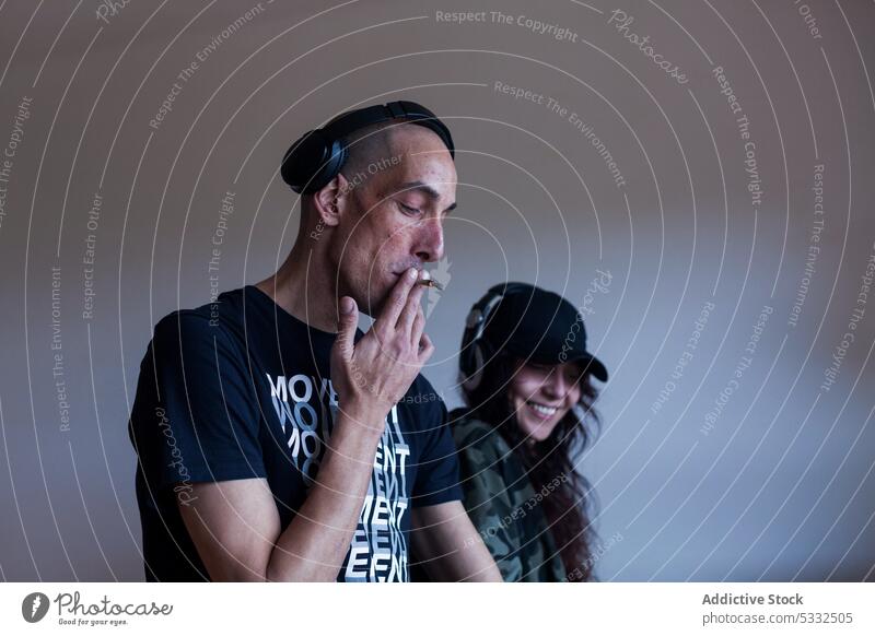 Male and female DJs in headphones in room man woman couple smile happy dj cigarette smoke listen modern cap music outfit girlfriend studio cool bald together