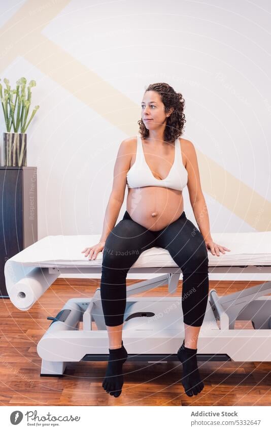 Smiling pregnant woman sitting in sportswear on massage table patient bench smile wait clinic relax healthy expect female belly happy tummy wellness maternal