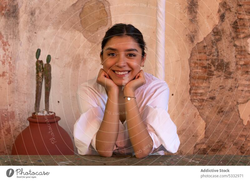 Happy woman sitting by wooden table outdoors smile happy cheerful touch chin positive plant dreamy green glad young san miguel de allende casual leisure
