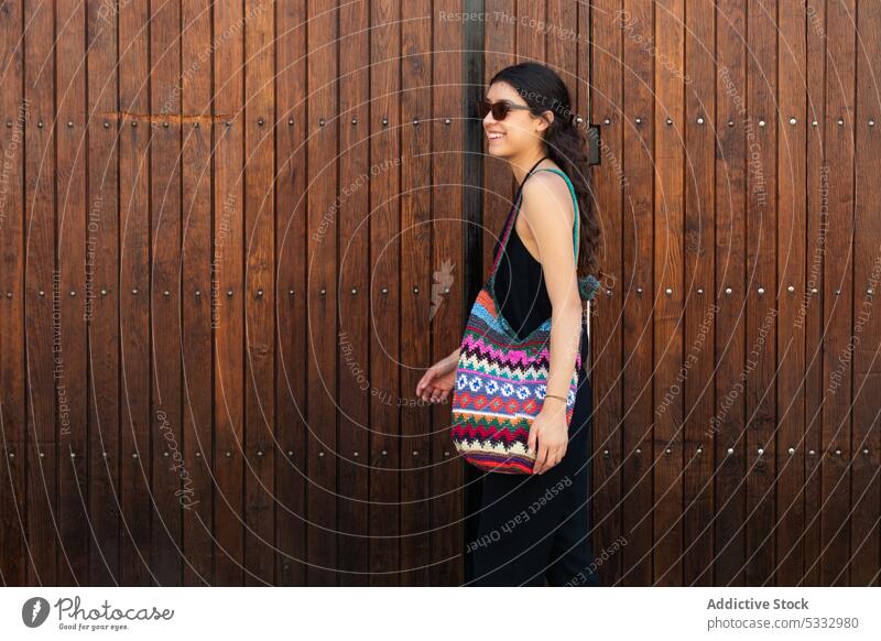 Young woman with colorful handbag standing by wooden wall trendy sunglasses model fashion smile cheerful positive happy female style confident black dress young