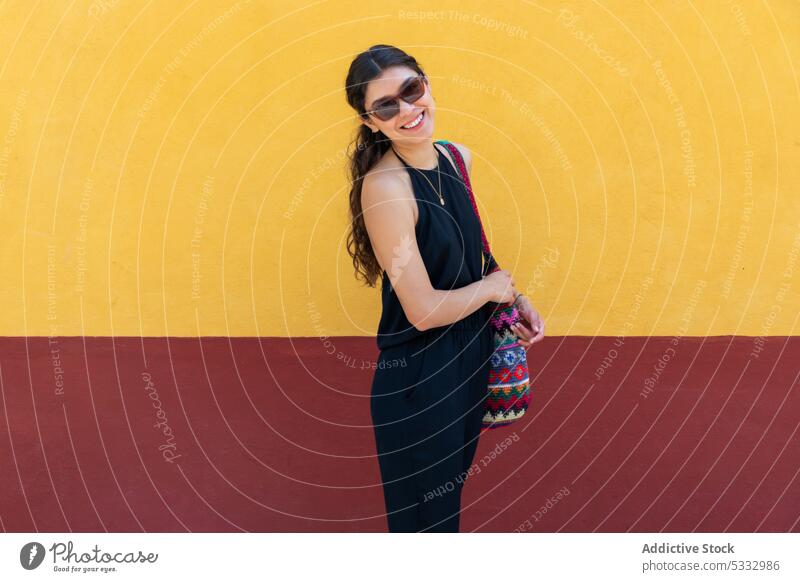 Smiling woman with handbag standing by colorful background sunglasses fashion wall positive style cheerful smile trendy bright outfit black dress apparel model