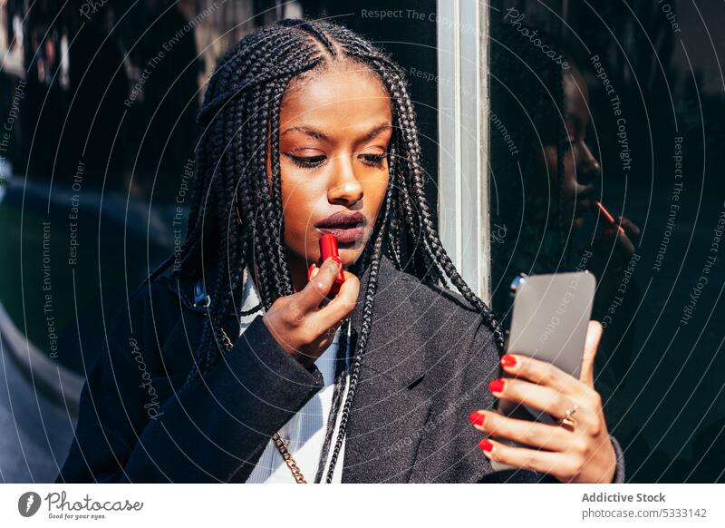 Black woman applying lipstick on street smartphone makeup using braid device mobile visage african american female ethnic black afro feminine appearance