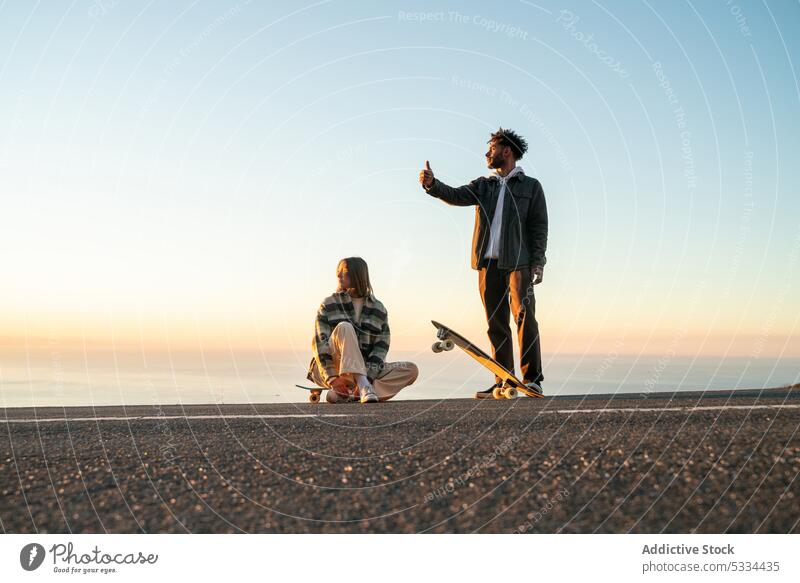 Happy multiethnic couple with skateboards having fun together on road smile sunset thumb up hitchhike gesture relationship love road trip happy skater seaside