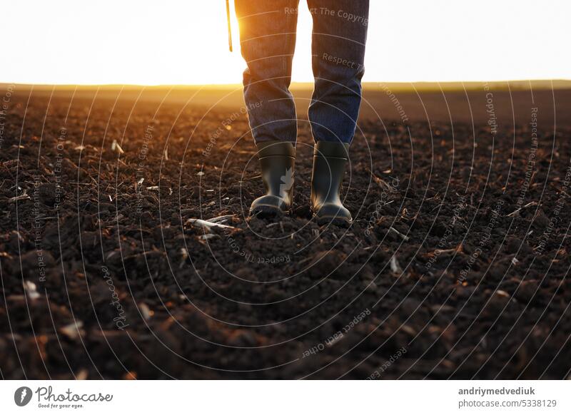Agriculture. Cropped shot of view businessman farmer in rubber boots walks along plowed field with digital tablet. Agronomist checking and analyses fertile soil on sunrise. Agribusiness.