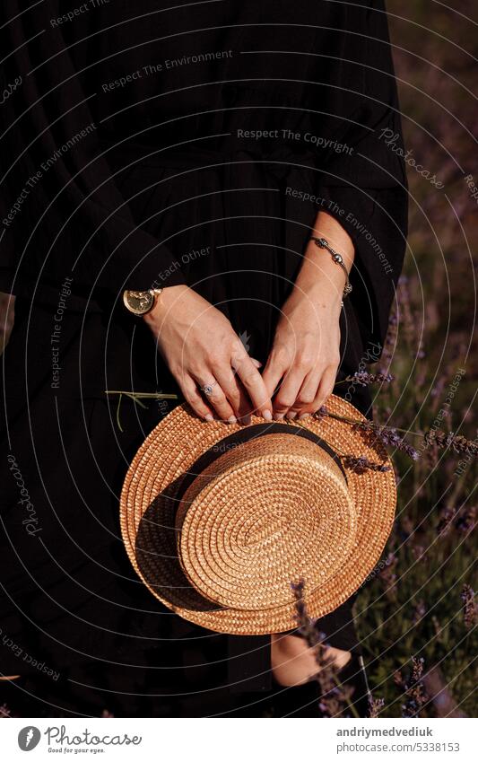 Cropped photo of woman in black dress holds straw hat and lavender flower in hands in blooming lavender field. Fashion beauty concept. Stylish girl with jewellery, watch on her hands. Women's day