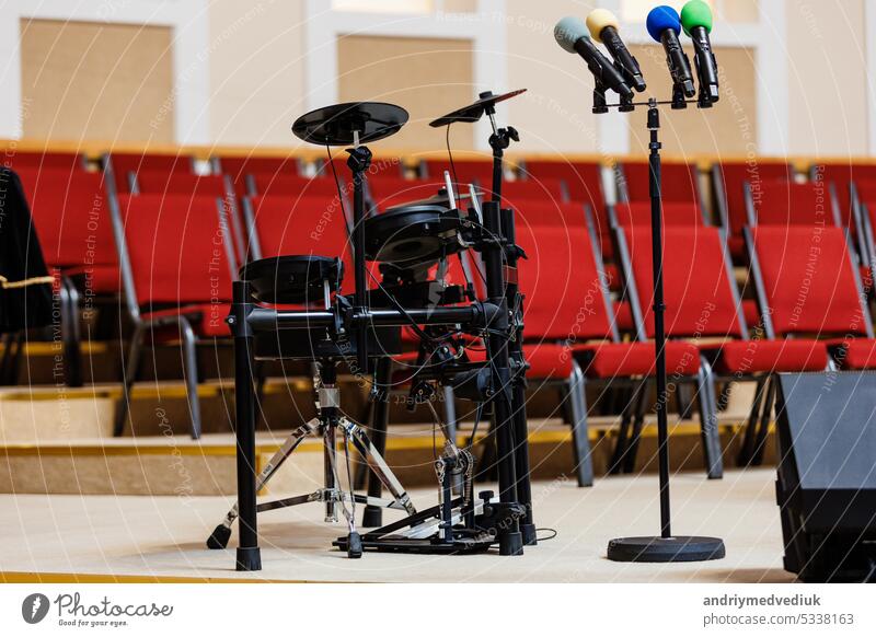 Coloured microphones on stand and electronic drum set on podium, a lot of red chairs blurred in the background. Concept of concert, event, communication, seminar, media, graduation, performance