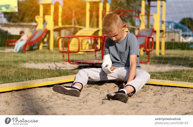 crying child with broken limb arm sits outdoors and looking how kid girl is playing on playground. concept of health accident, and medical. the worst summer vacation.