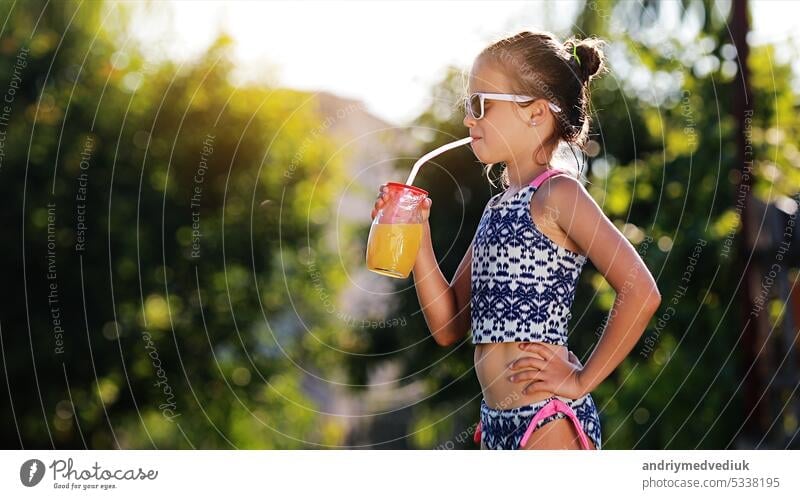Beautiful little girl, cute toddler with sunglasses is drinking orange juice and shows thumb up. Kid in in swimwear having fun during family vacation in a resort near pool. summer holidays
