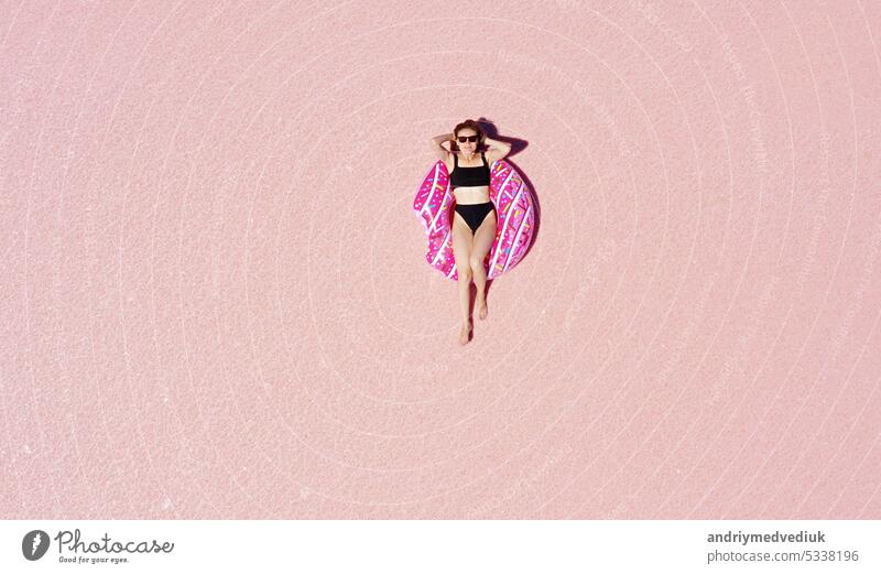 World Tourism Day. Above view of young happy woman in sunglasses and black bikini sunbathing lying on an inflatable pink donut in a sunny pink lake. Enjoying holiday vacations. Place for advertising