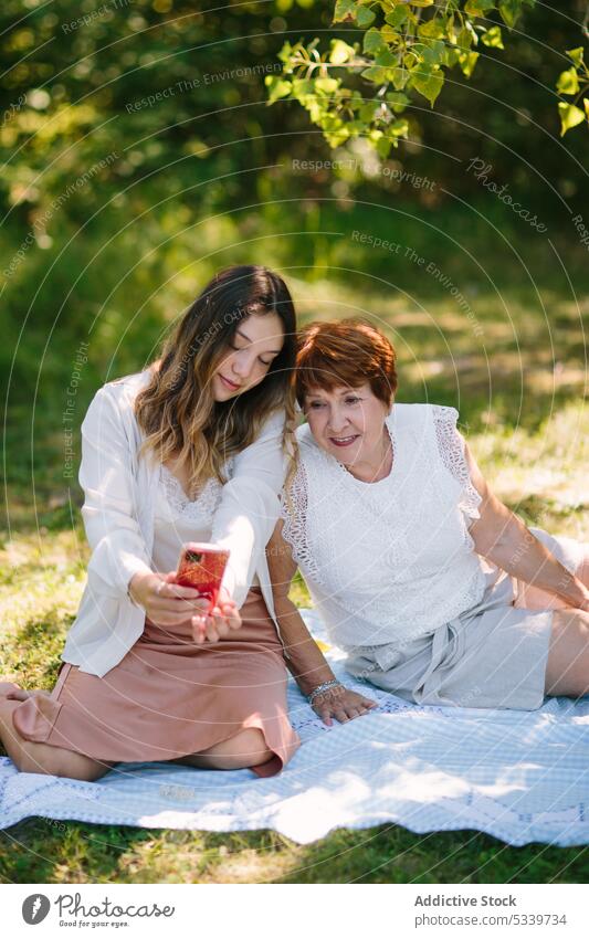 Content teen girl with smartphone near senior woman on picnic blanket granddaughter together share park grandmother social media using teenage relationship