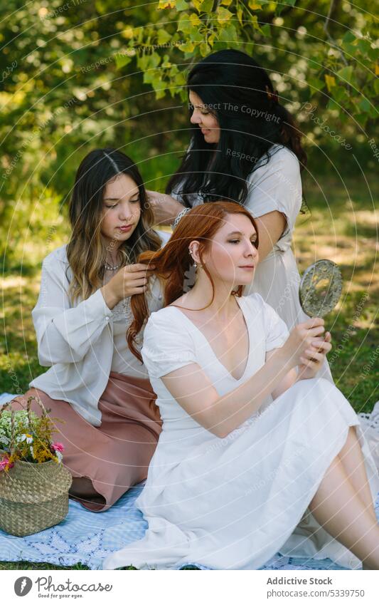 Calm women doing hairstyles to each other in park picnic summer friend together lawn meadow grass friendship blanket nature weekend happy sunlight carefree